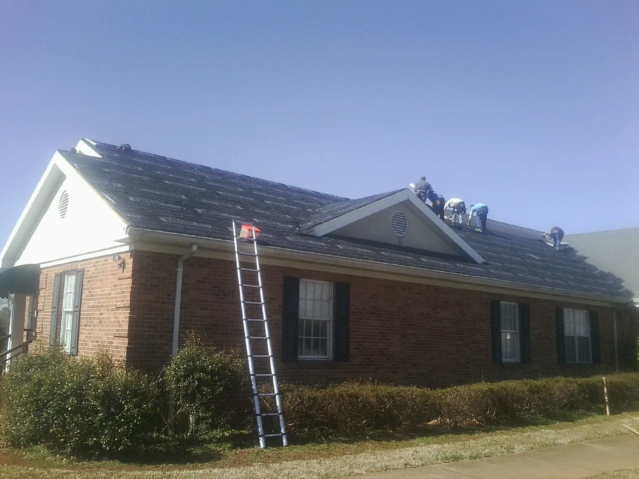 Crew tearing shingles off a brick ranch roof with a ladder set — Metal Roof Repair work by our crew in Lake Panasoffkee