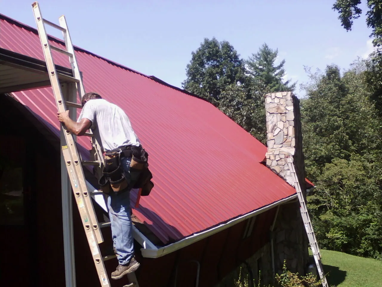 Roofer on a ladder beside a red metal roof and stone chimney — Soffit & Fascia Repair by Roofing Brothers in Lake Panasoffkee